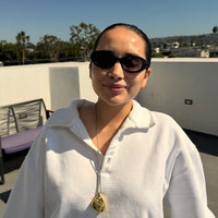 A woman in a white polo shirt and sunglasses on a sunny rooftop terrace, holding a brown leather bag