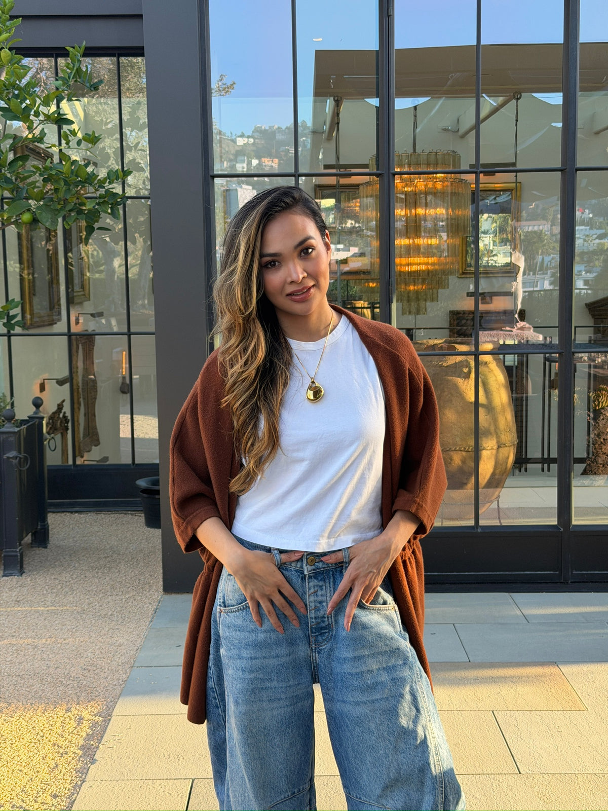 Woman wearing gold Legend One AI memory pendant with white t-shirt and jeans, standing outdoors in front of glass building, full-body lifestyle photo
